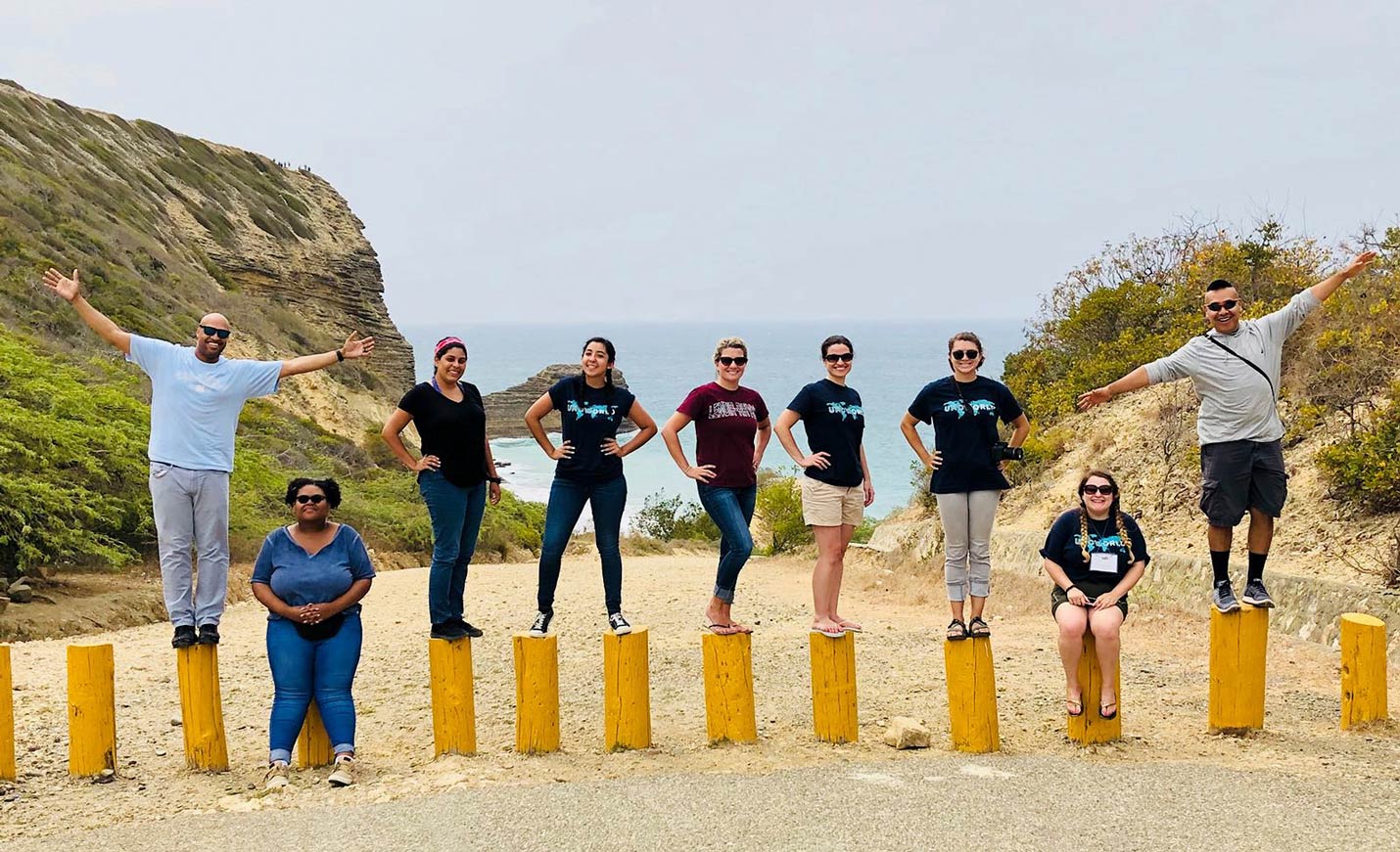 A group of students, some wearing UNCW World T-shirts, stand on pilings on a rocky beach