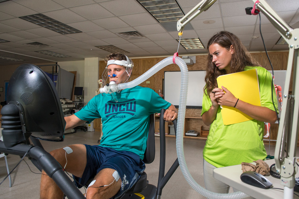 One student sits with breathing gauges on face while another stands and studies the readings.