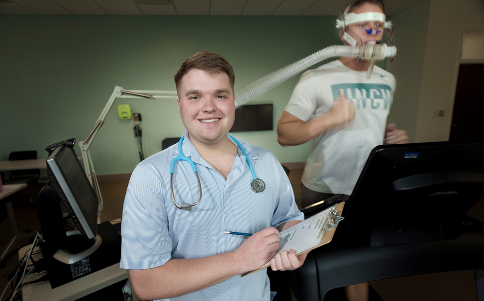 Nolan Byrd holds a clipboard and a pen and stands in front of a male on a treadmill who has a tube in his mouth.