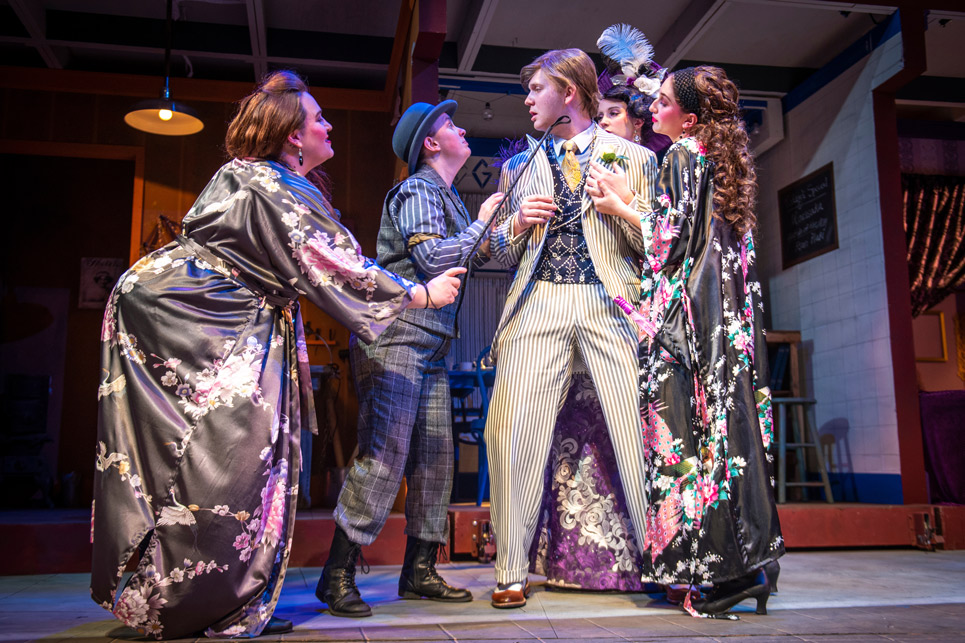 People on stage surround main character in an old fashioned play.