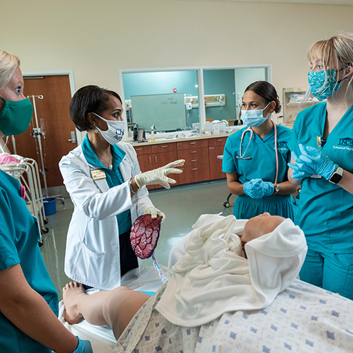 Nursing professor and three students stand around a practice mannequin in a bed