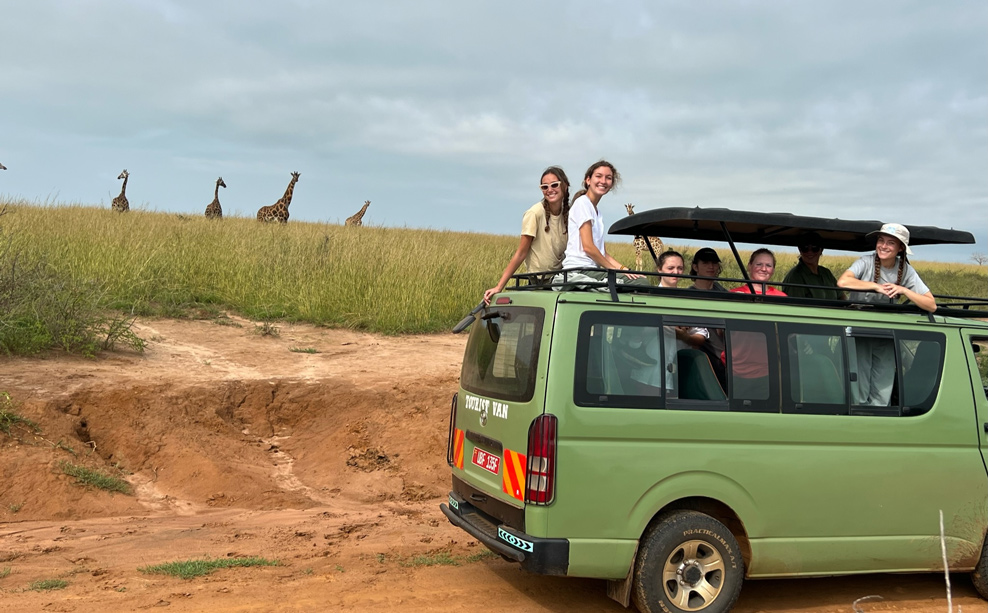 Students sit atop a green bus with giraffe in the distance.