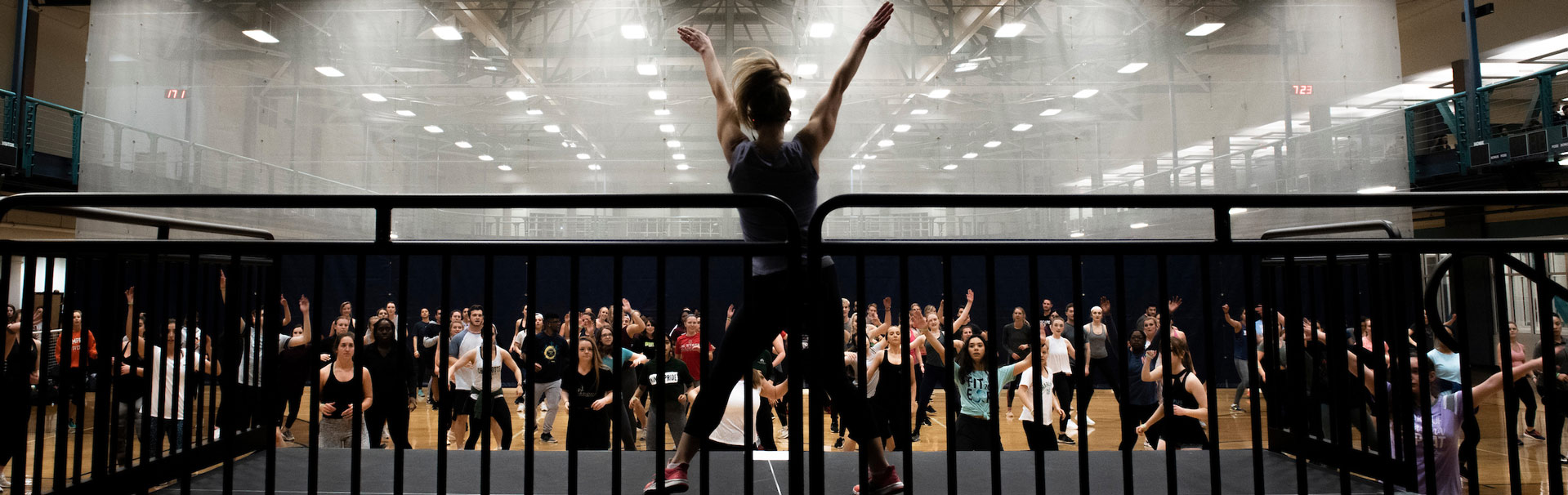 A fitness instructor is on a platform, instructing a gym full of participants. Her arms are above her head in a V formation.
