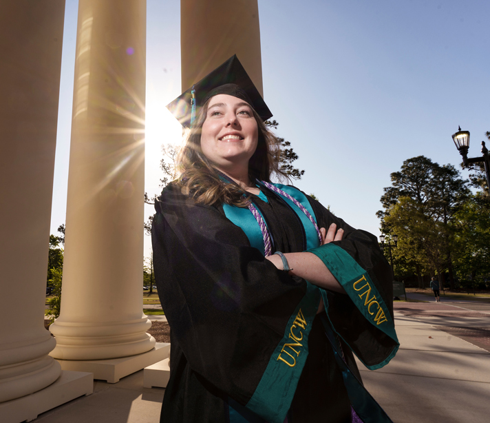 Maddie Picken, wearning a black graduation cap and gown with a teal stole and purple cord around her neck, stands in front of three tall columns with her arms crossed and has a big smile on her face.