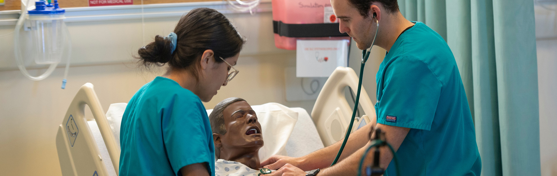 Two nursing students in scrubs examine a medical dummy in the Simulation Lab