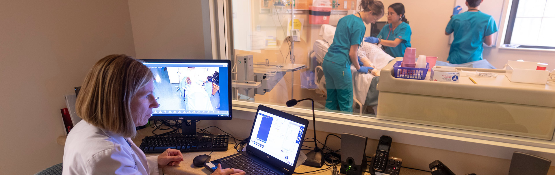 A female School of Nursing faculty looks at a laptop and larger screen. She is observing three nursing students through a large window in the simulation learning center as they perform tasks on a mannikin. 