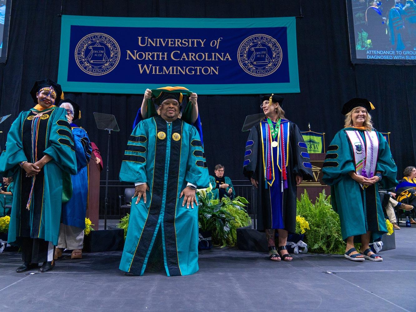 Four DNP students wearing commencement robes and caps stand on the stage to receive their doctoral hoods.