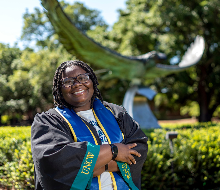 Nakyah McNeal stands in her graduation robe in front of a large Seahawk sculpture on the UNCW campus.