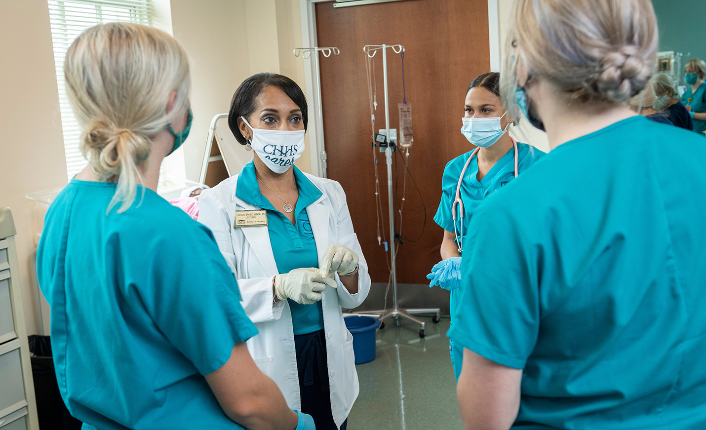 A nursing professor stands with three nursing students forming a semi-circle around her as she lectures to them.
