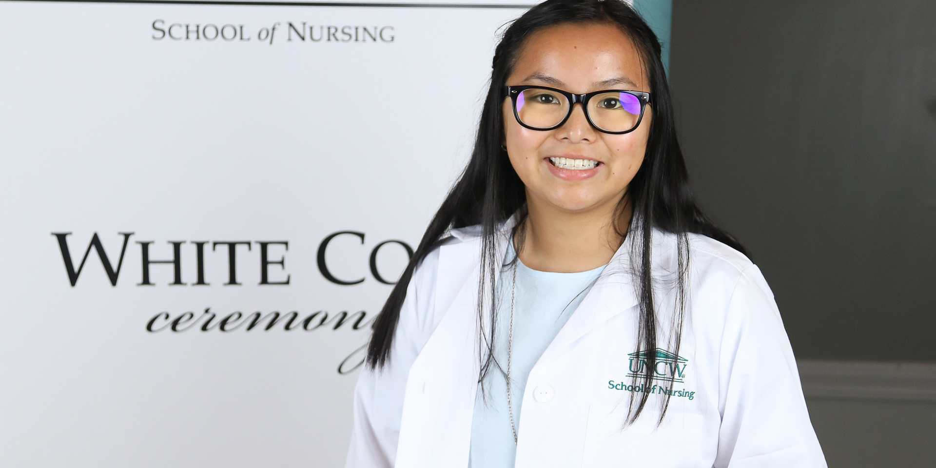 A nursing student stands in her white coat in front of the sign for the white coat ceremony.