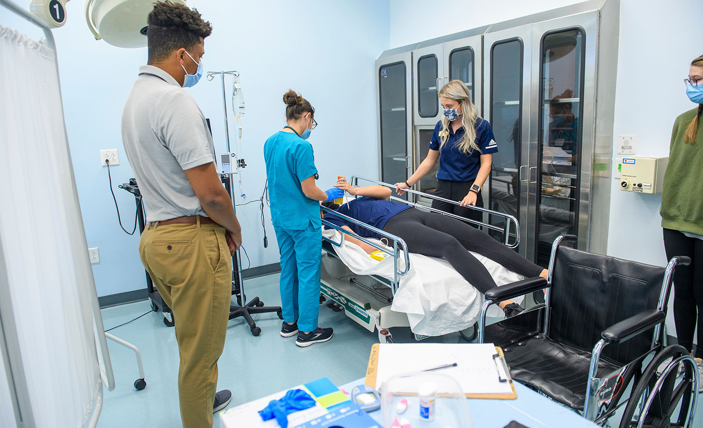 A nursing student and athletic training student work together to tend to a patient on a hospital bed in a mock treatment session.