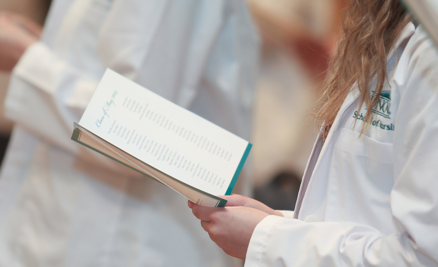 The hands of a nursing student holding the program while wearing their white coat during the White Coat Ceremony.