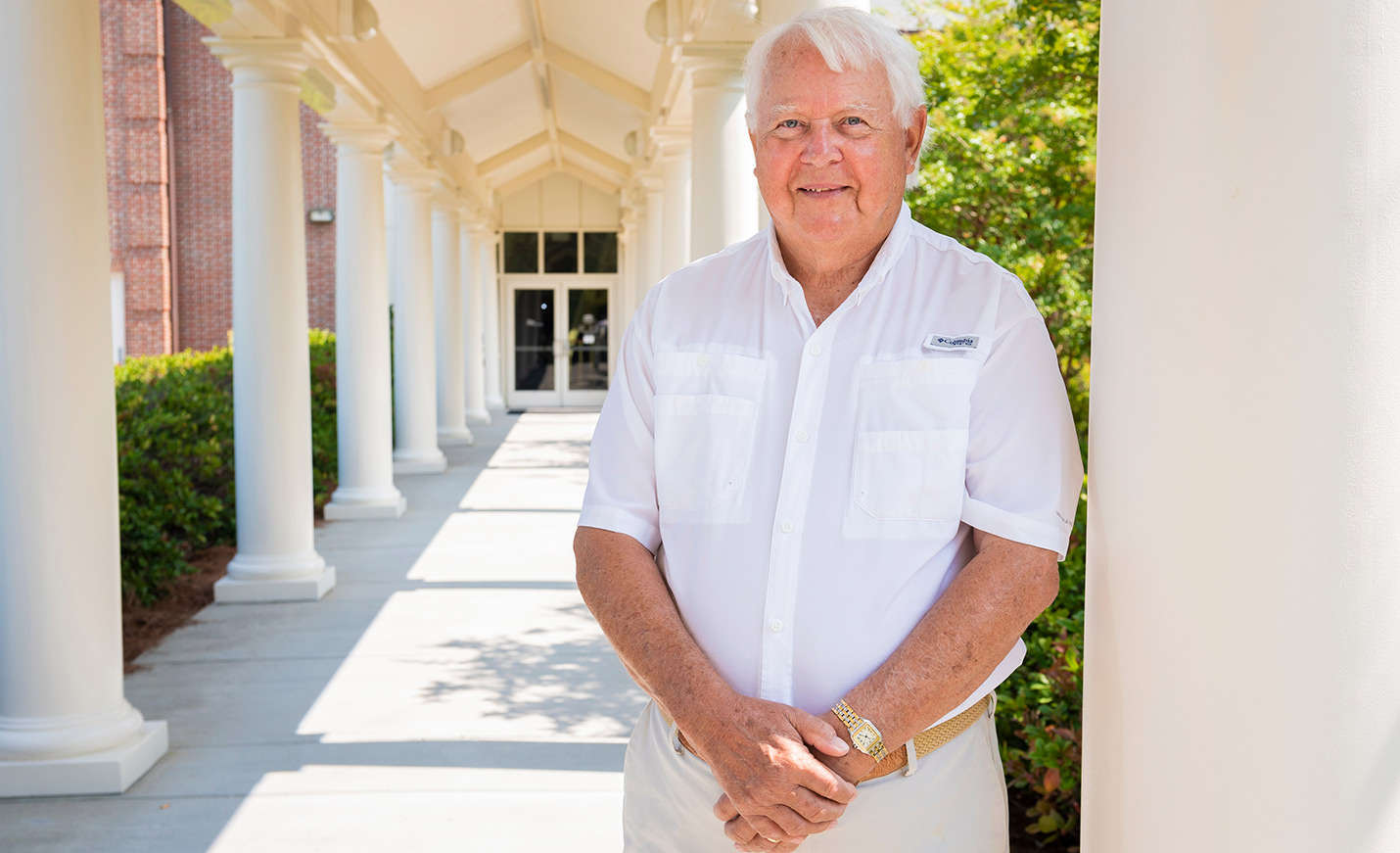 An older adult man stands with his hands clasped while leaning against white pillars on UNCW's main campus.