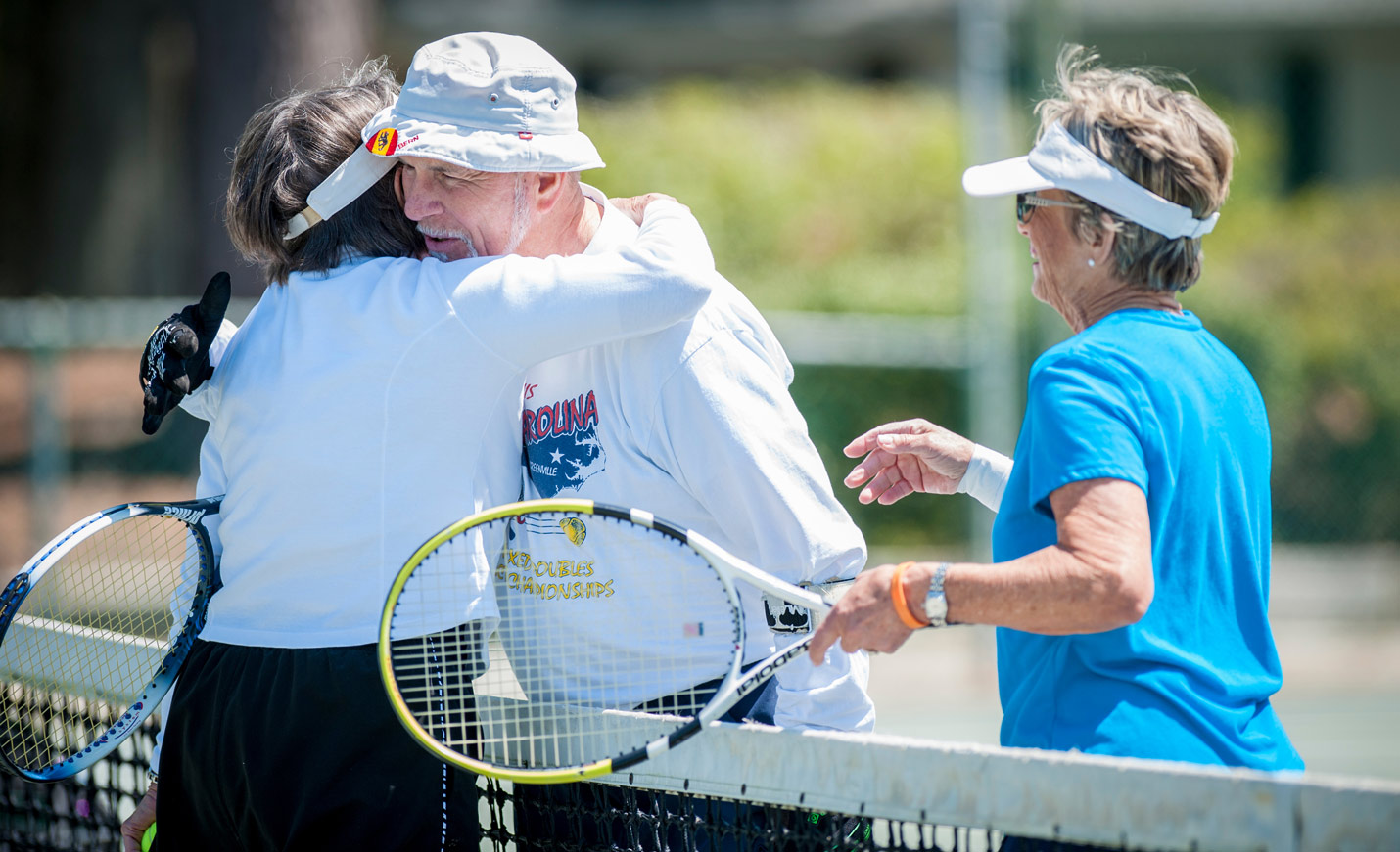 Two older adults hug over a tennis court net after a game.