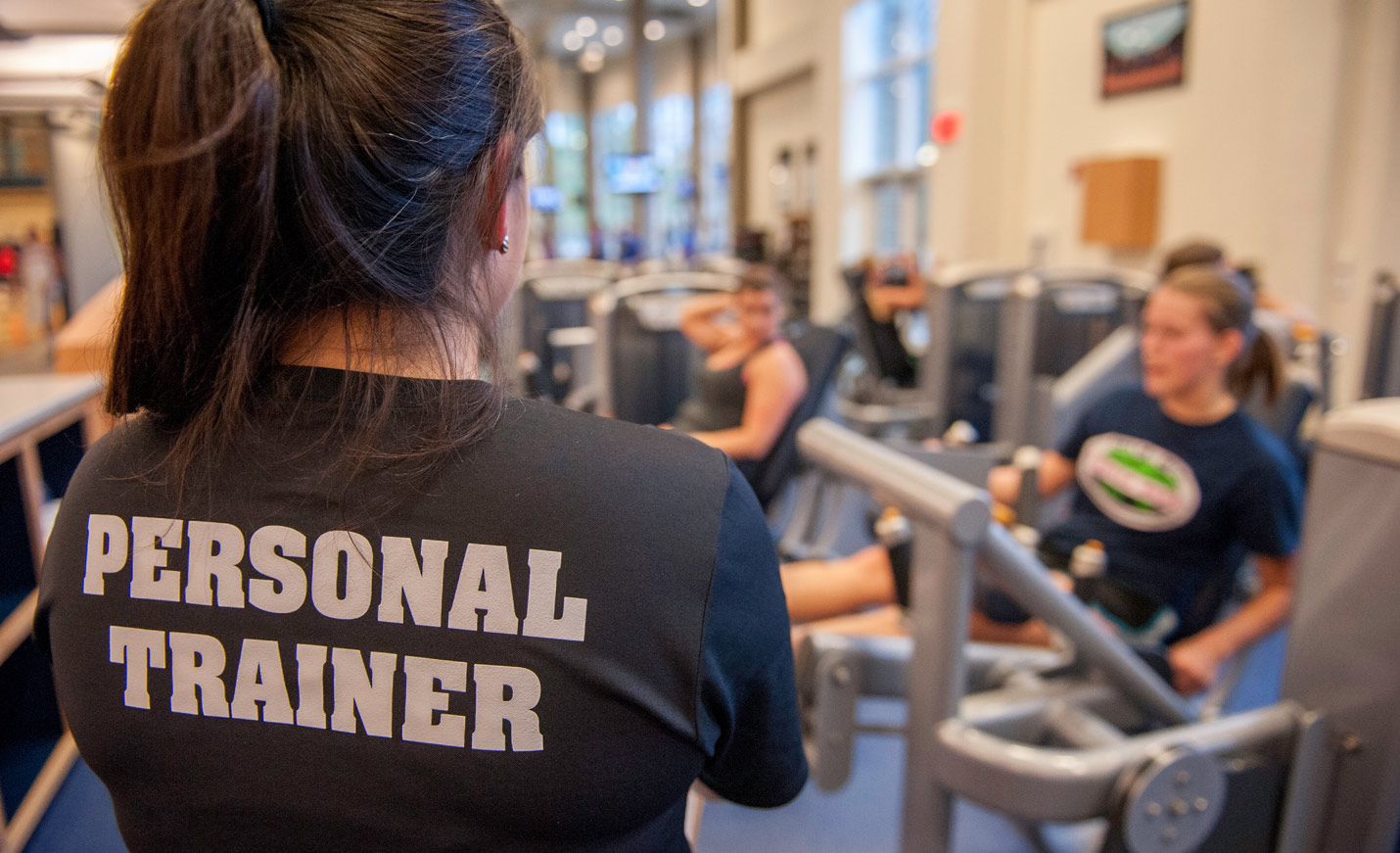 A woman stands with her back to the camera, her shirt reads personal trainer. She is overseeing students exercising.