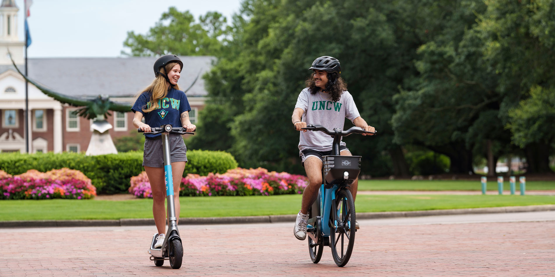 Two students in UNCW T-shirts ride e-bikes on campus.