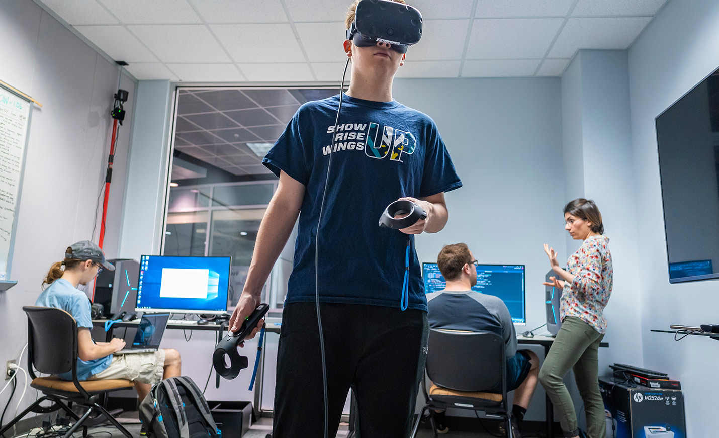 A student stands while wearing VR headset; two other students are seated at computers in the background