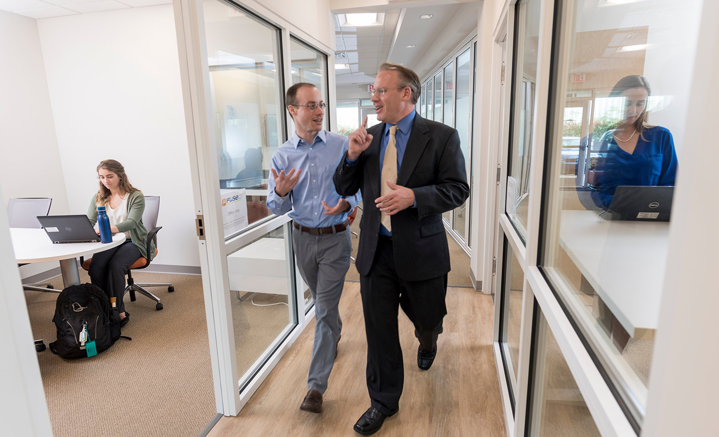 Two men walk down a hallway surrounded by glass-walled offices