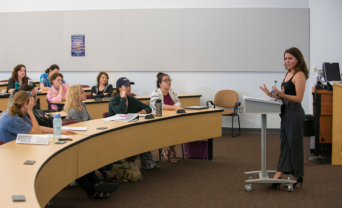 A faculty member talks in front of a group of students seated at tables in a lecture hall