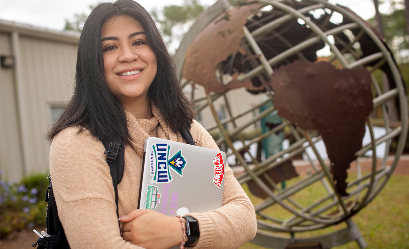 A student stands in front of a globe statue and holds a laptop
