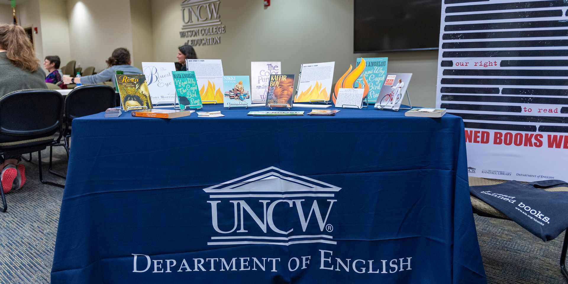 A picture of a desk with books on it and a tablecloth that reads, “UNCW Department of English”