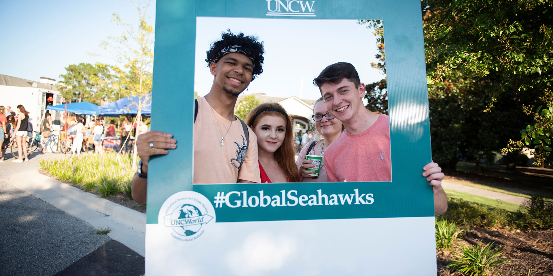 A sign frames three students and says “#GlobalSeahawks”