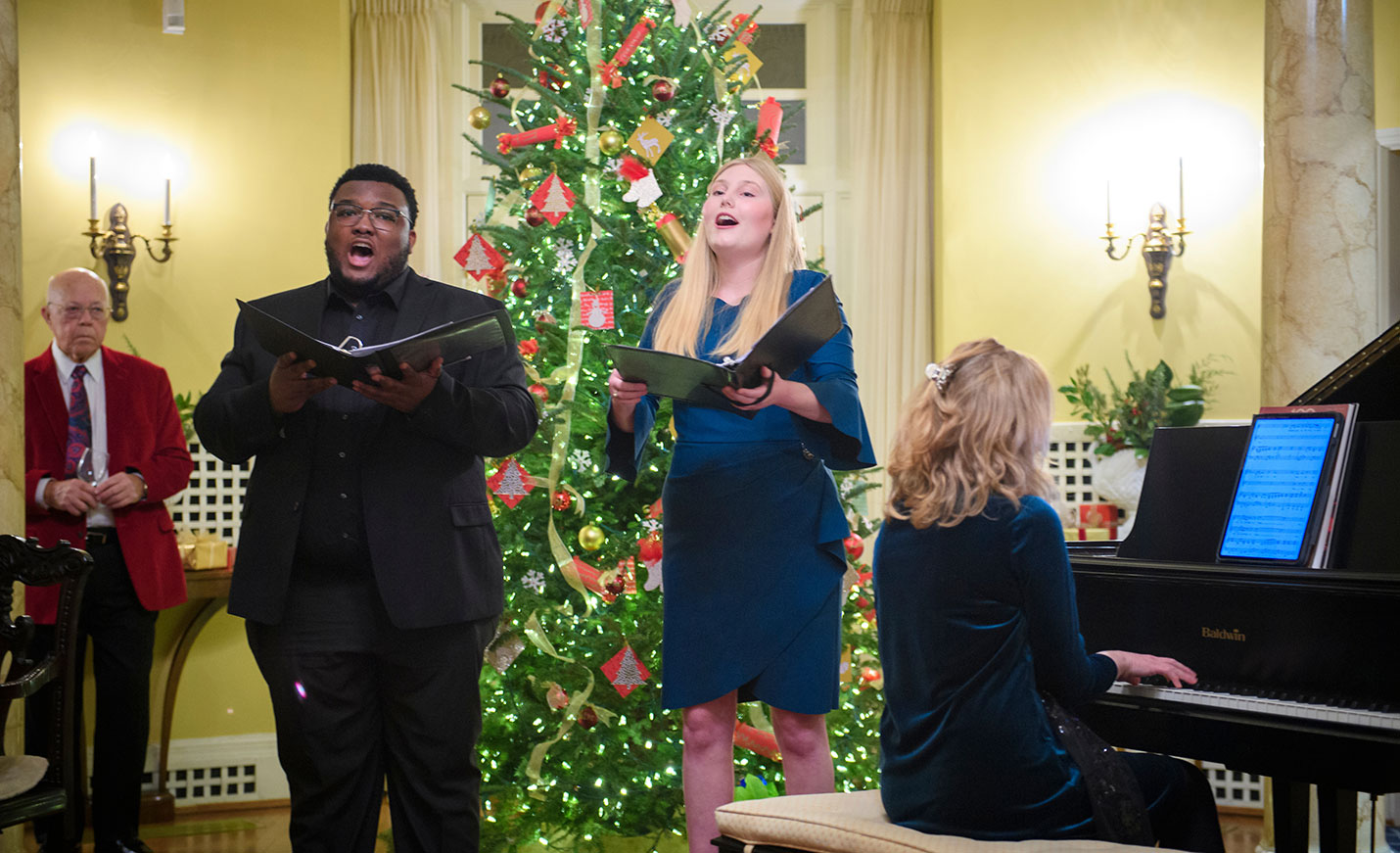 Two students singing and one playing piano in front of a holiday tree at Kenan House