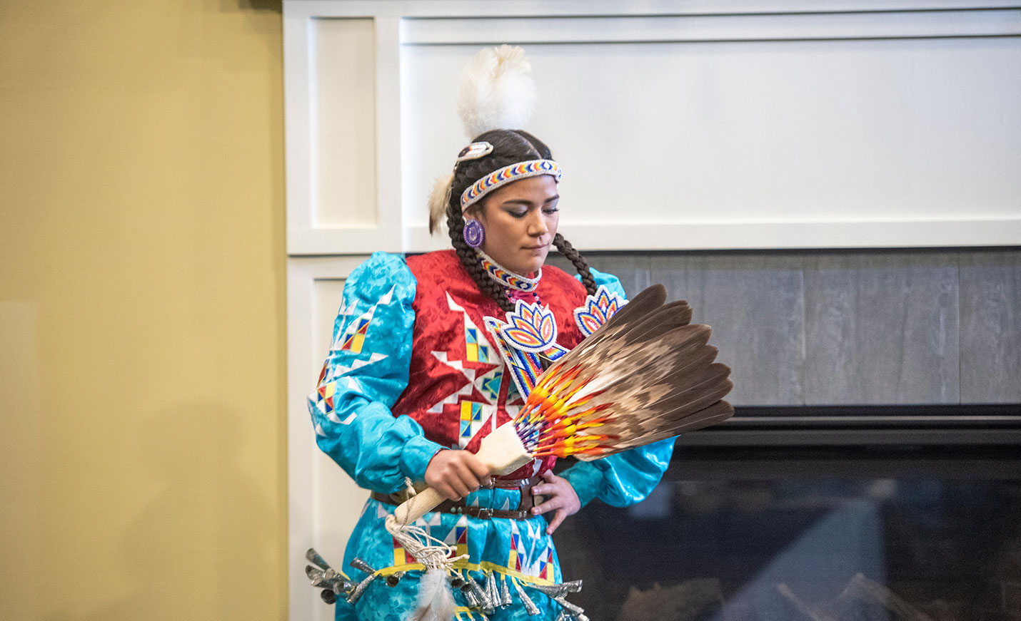 A woman in colorful Native American attire holds a bouquet of feathers while performing a dance