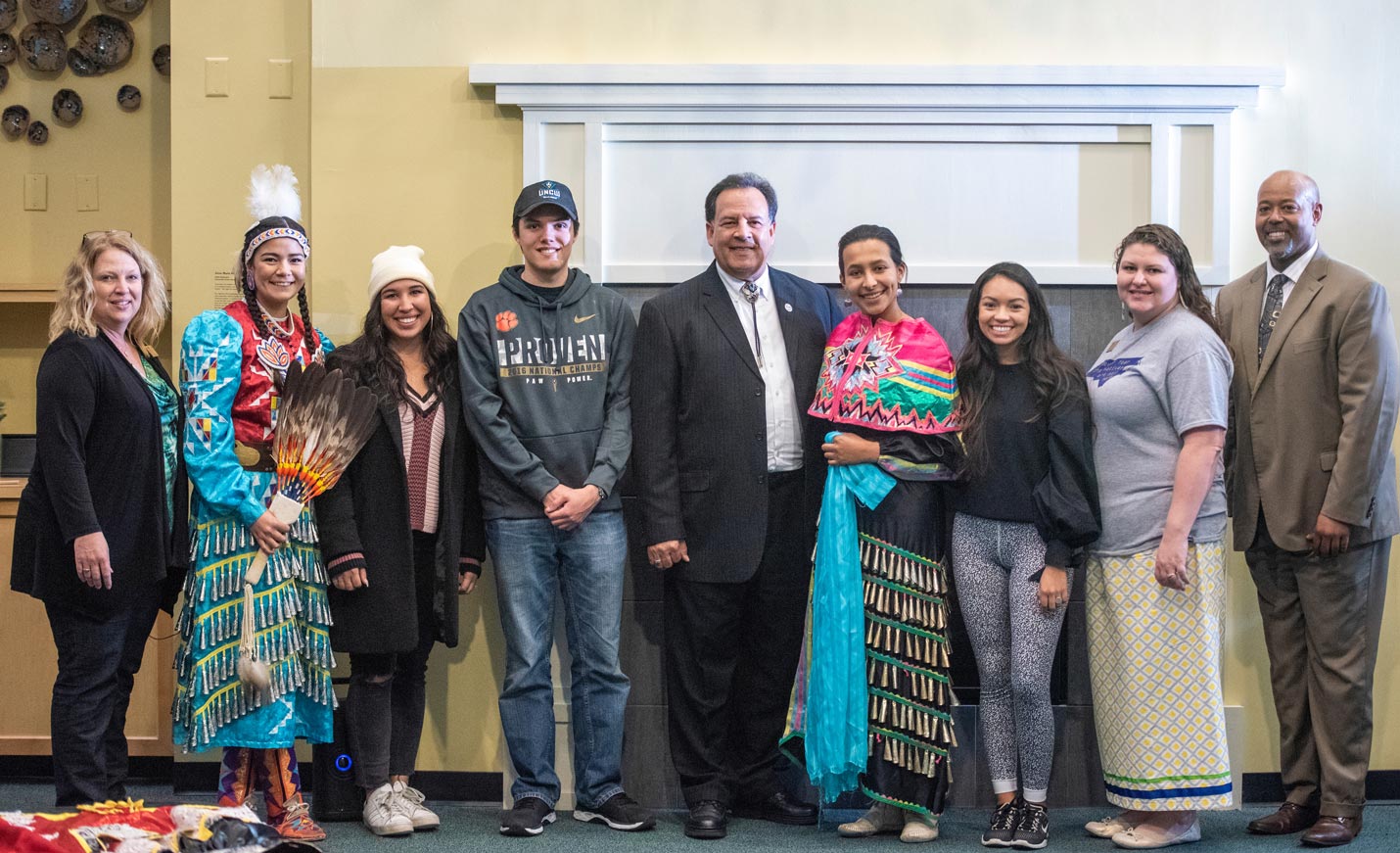 A group photograph of nine people at a Native American Heritage celebration. Two women are in colorful Native American attire. the man in the middle is wearing a bolo tie with his suit.