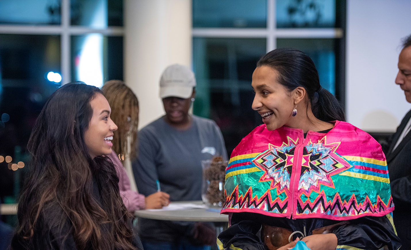A woman in colorful Native American dress talks with another woman. In the background are other students.