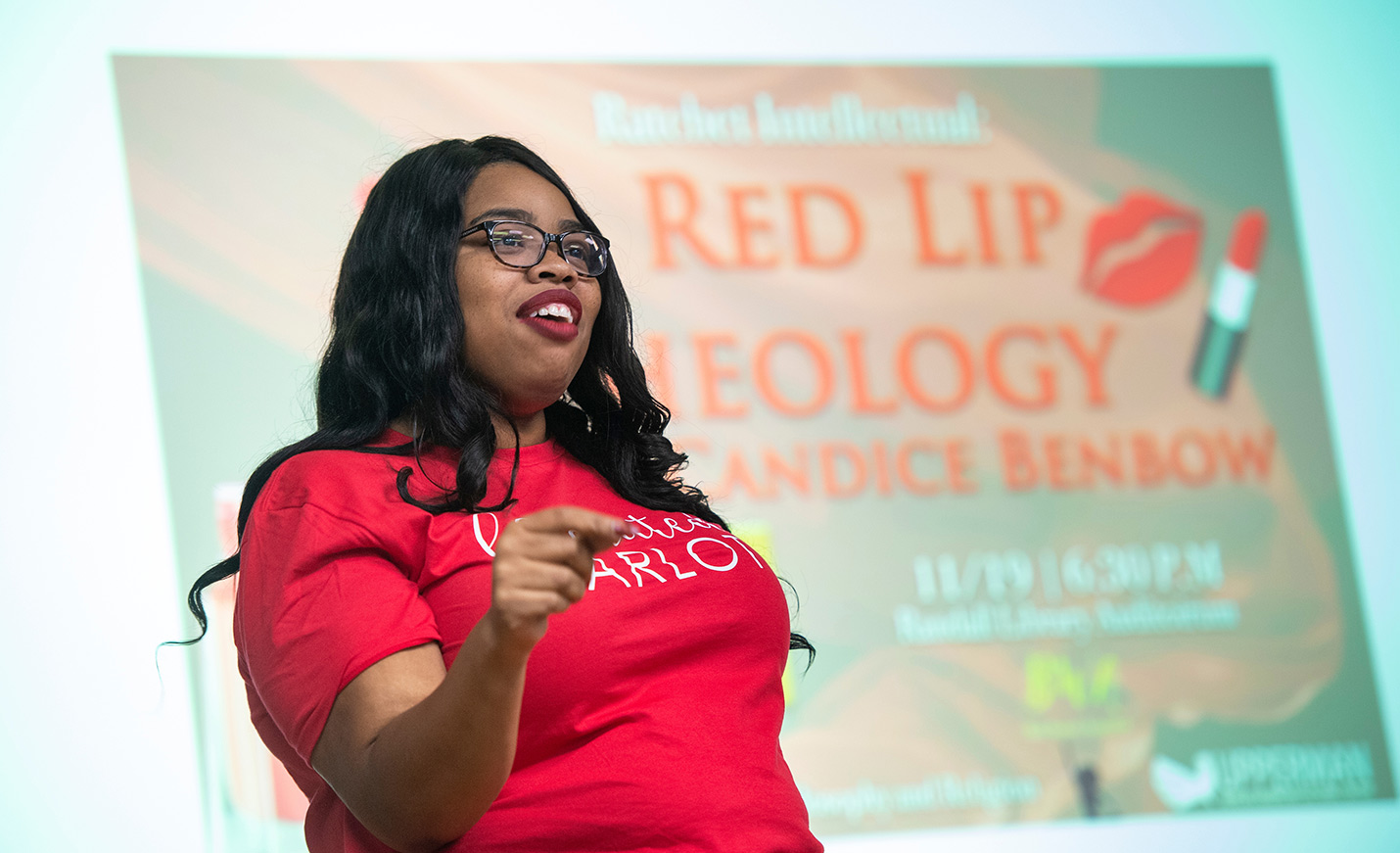 A woman in a red T-shirt with lettering stands in front of a screen with Red Lip Theology projected onto it