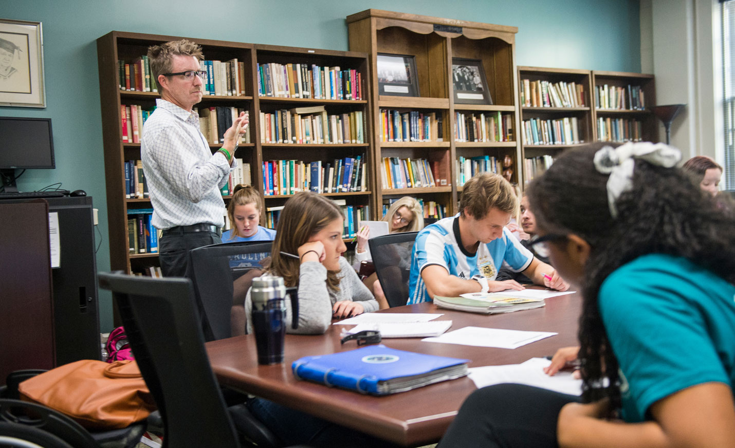 An instructor teaching a group of students seated at tables in front of shelves of books