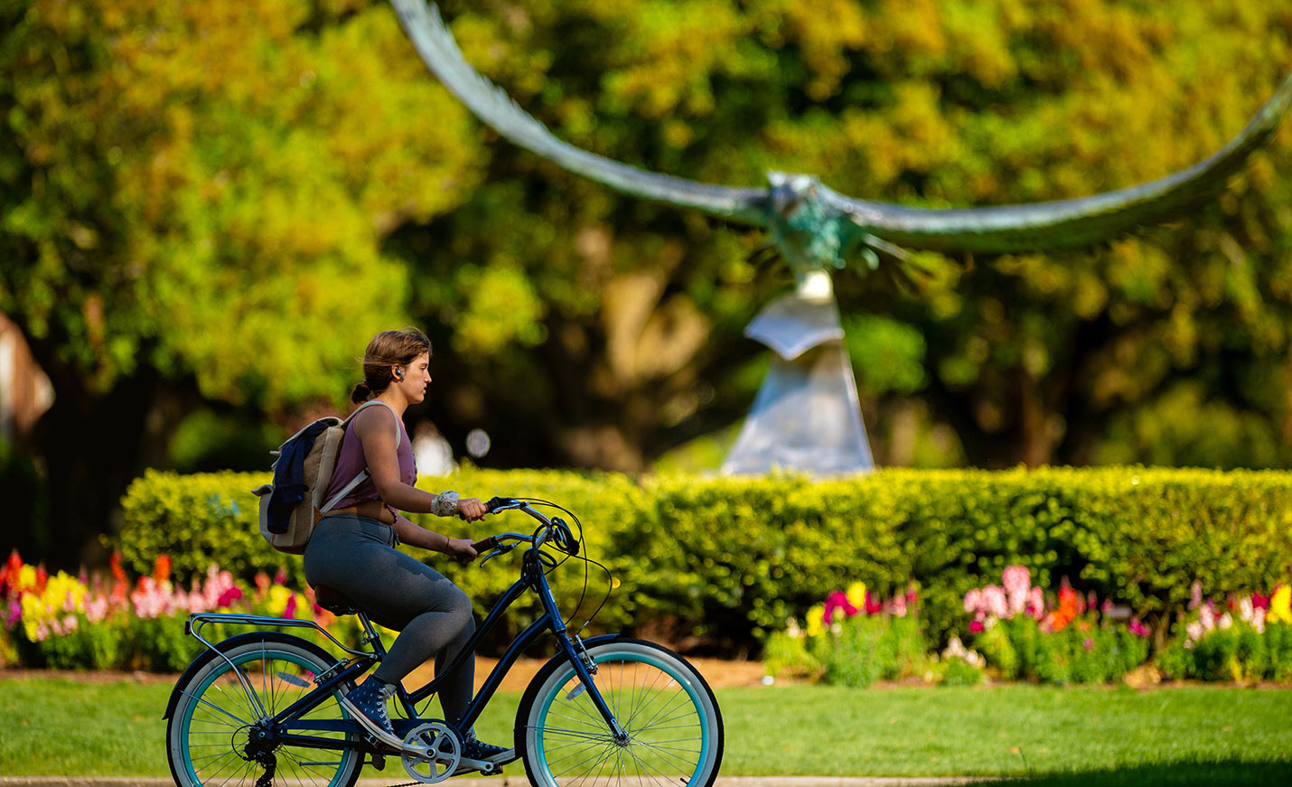 A student rides a bike past the soaring Seahawk sculpture. The background also includes colorful flowers and shrubs.