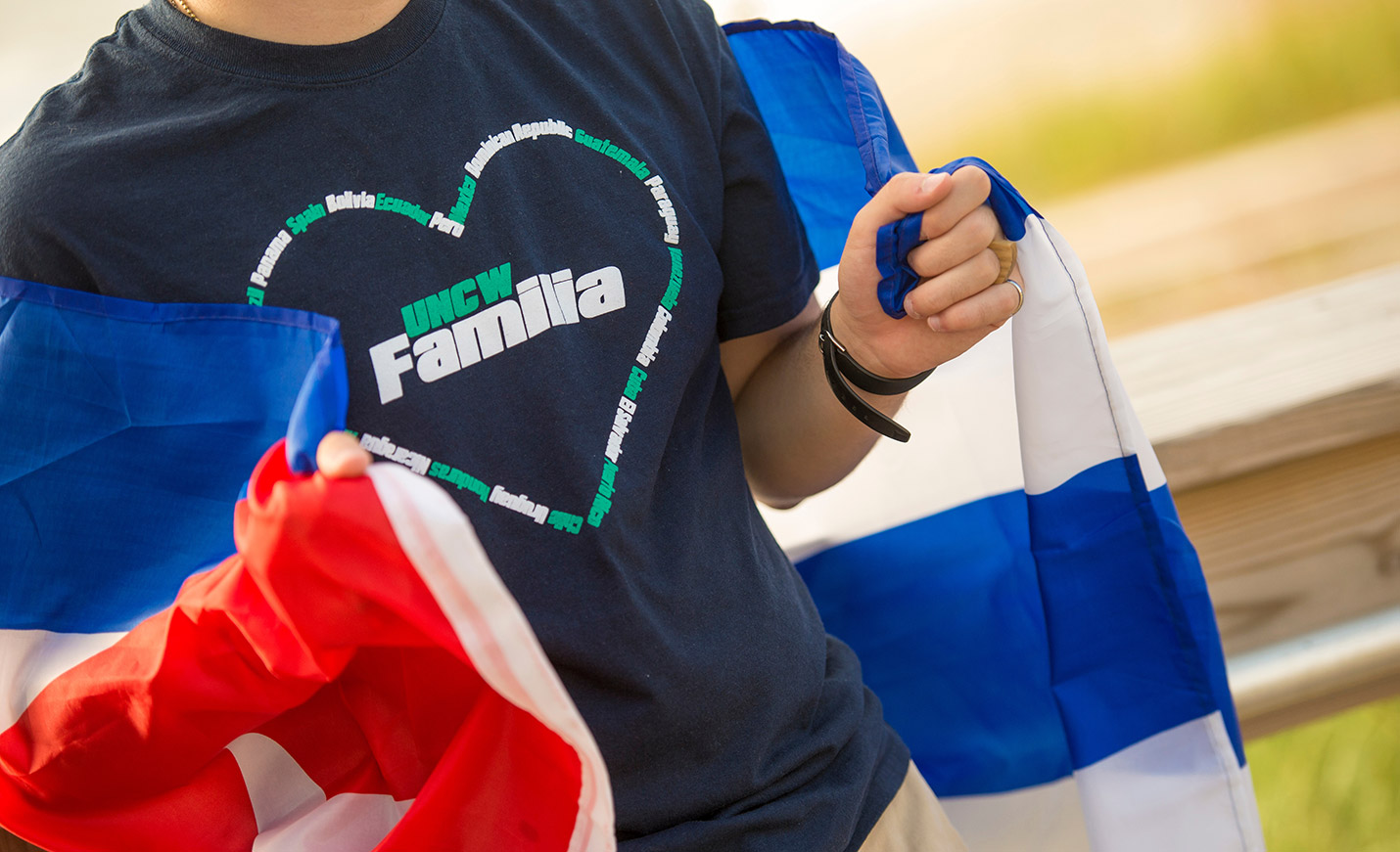 Close-up image of a person in a UNCW Familia T-shirt holding a blue and white flag