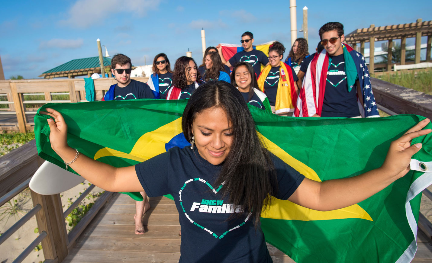 A young woman holding a Brazilian flag behind her and wearing a UNCW Familia T-shirt with other students in the background