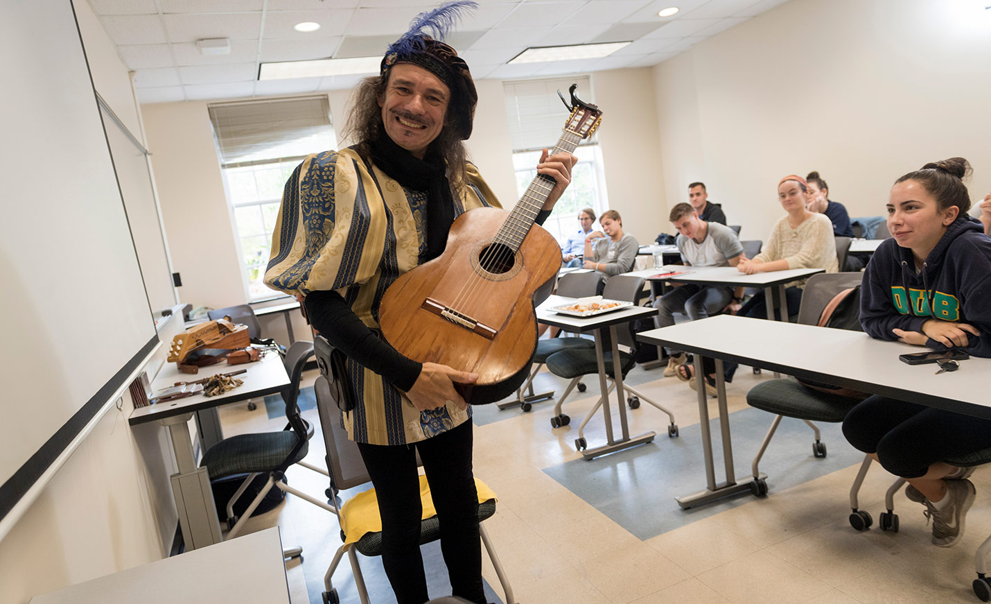 A man dressed as Don Quixote holds a guitar at the front of a classroom full of students