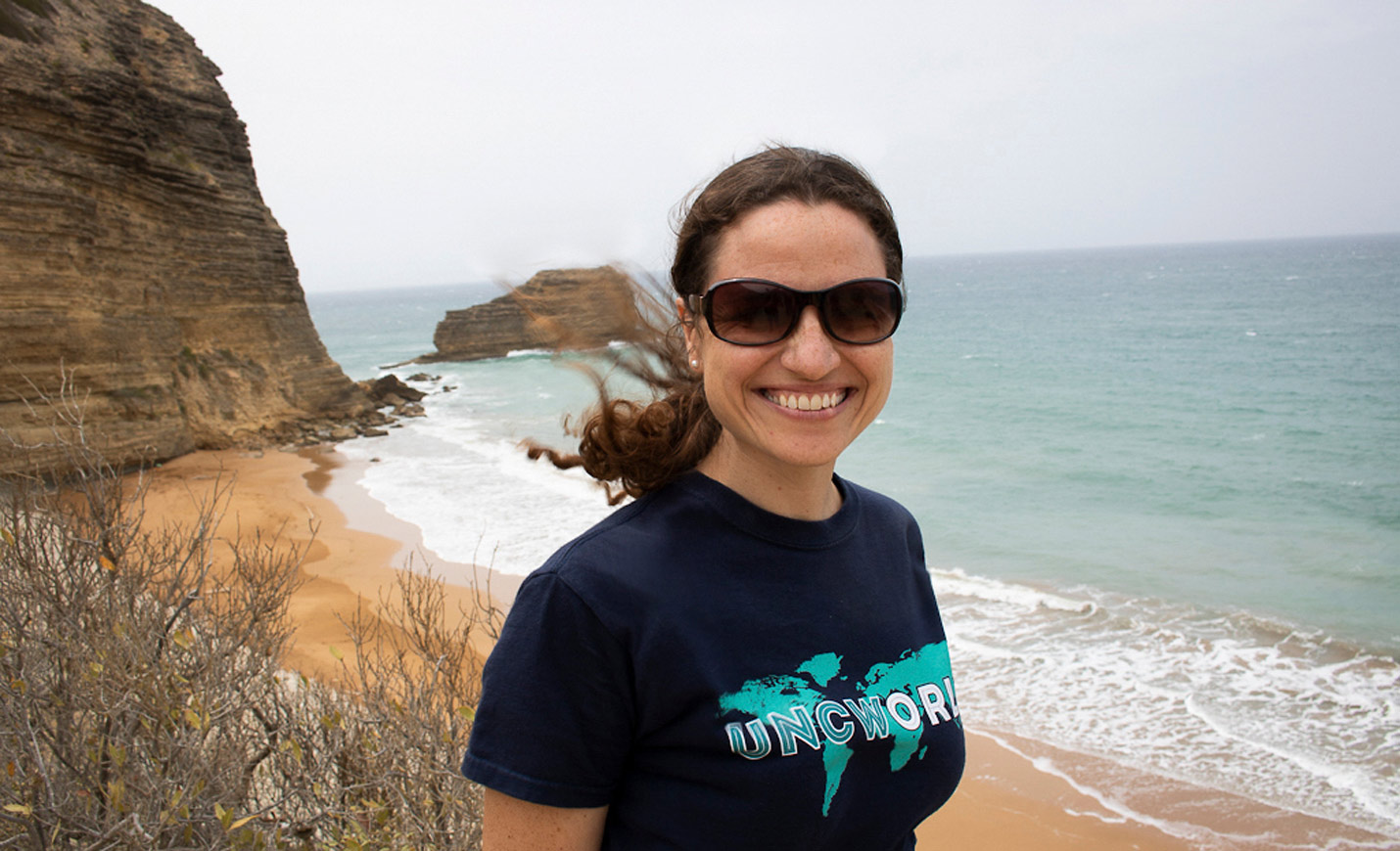 A woman wearing a UNCW World T-shirt poses on a rocky beach