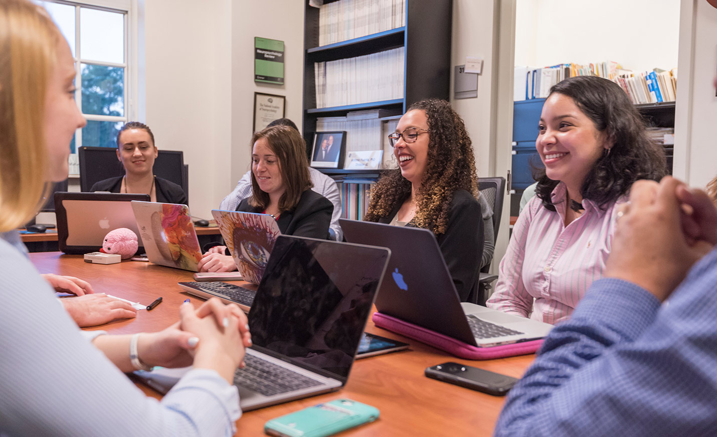 Smiling students hold a discussion while seated at table with laptops in front of them