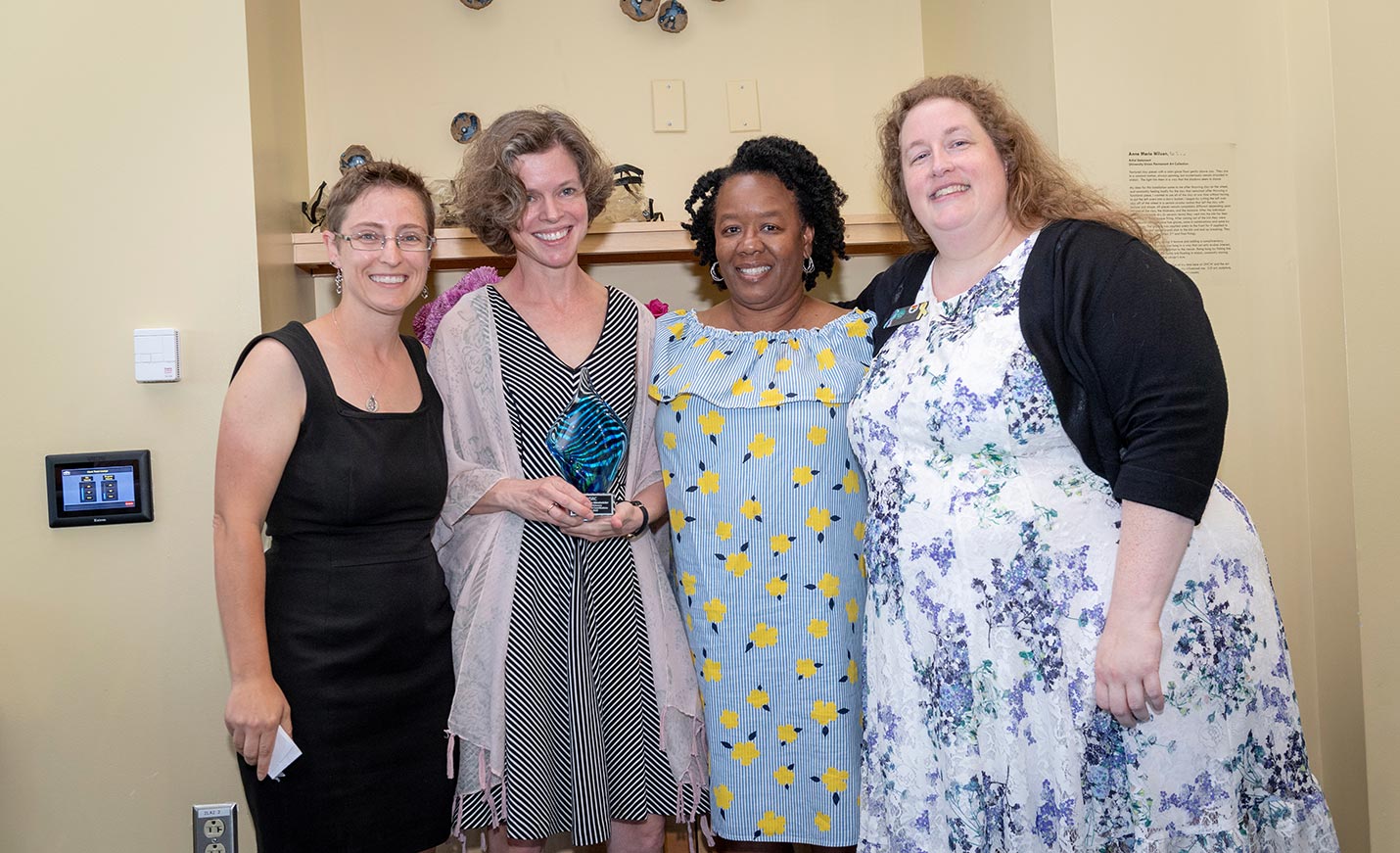 Four women stand together; the one second from left is holding a small award or trophy