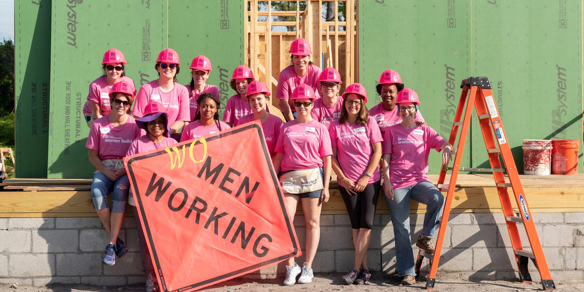 Group of people standing at construction site with construction sign that says “Wo” Men Working
