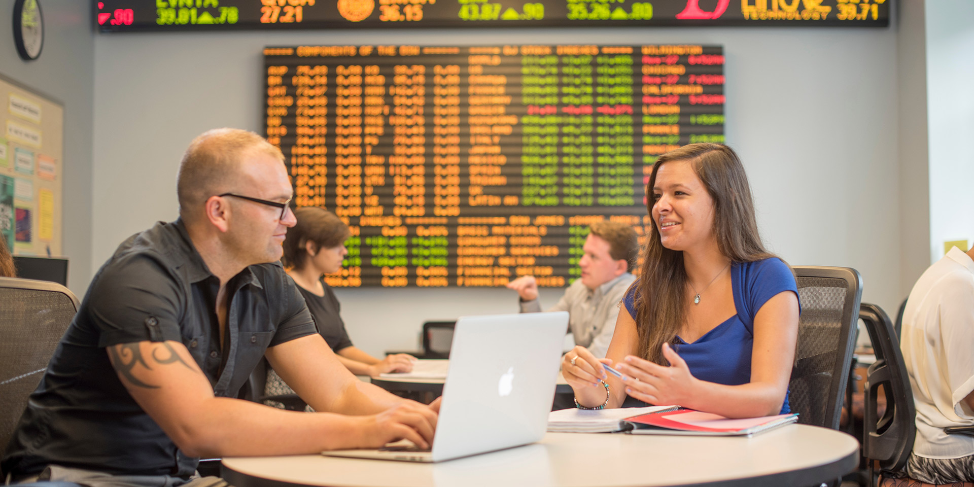Two students talk at a table in front of a large stock ticker board