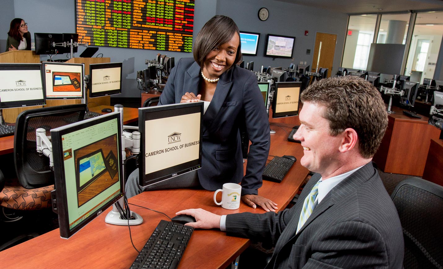 Two smiling people in a computer lab, one sitting on table and the other working on a computer screen