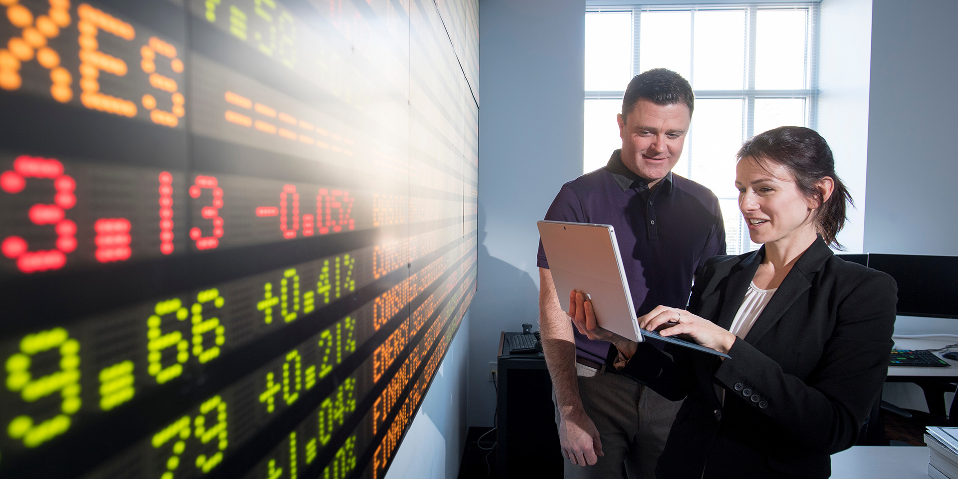 two people discuss something on a laptop as they stand in front of a large stock ticker board