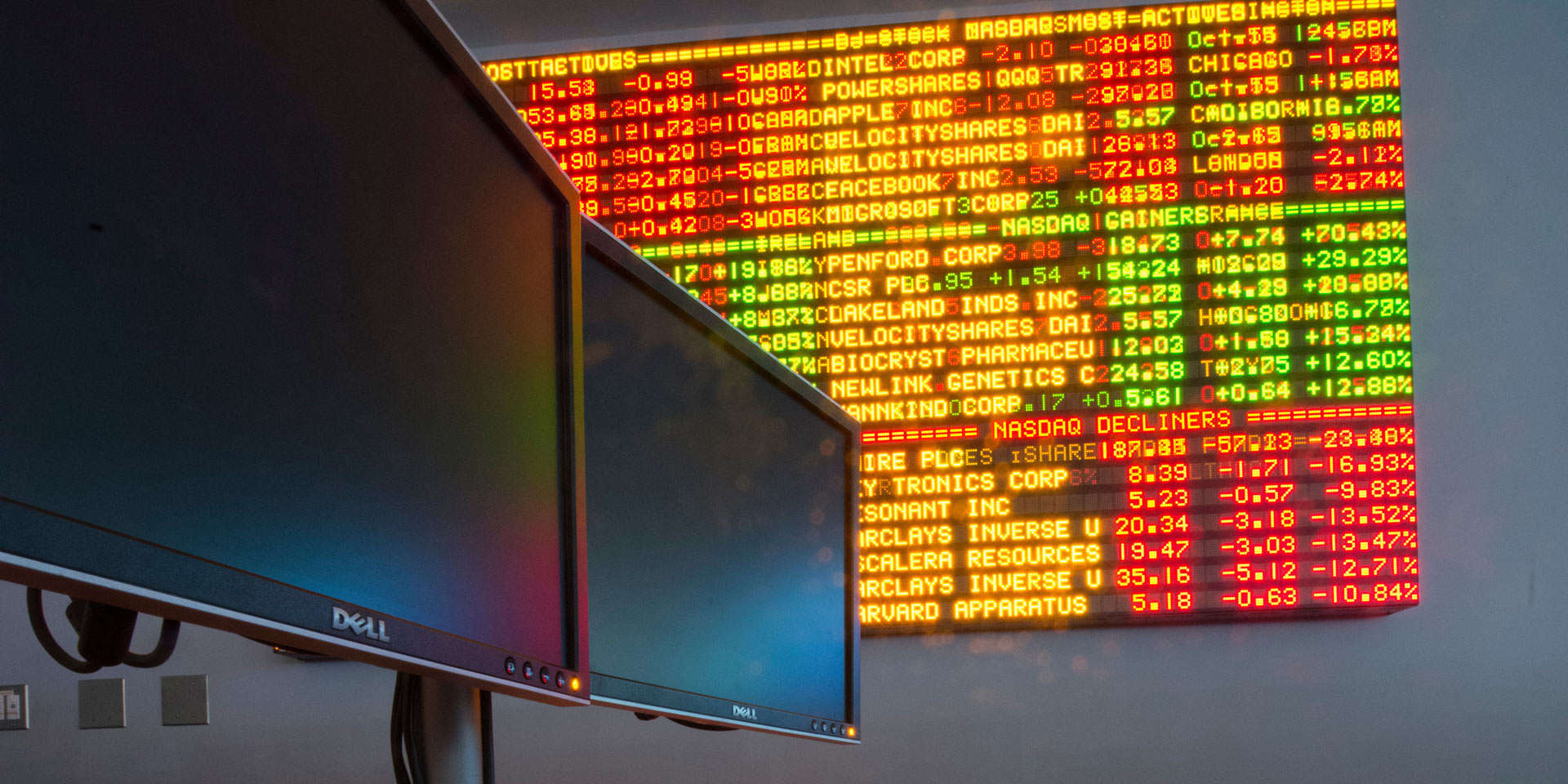 A closeup of two desktop monitors in front of a large stock ticker board