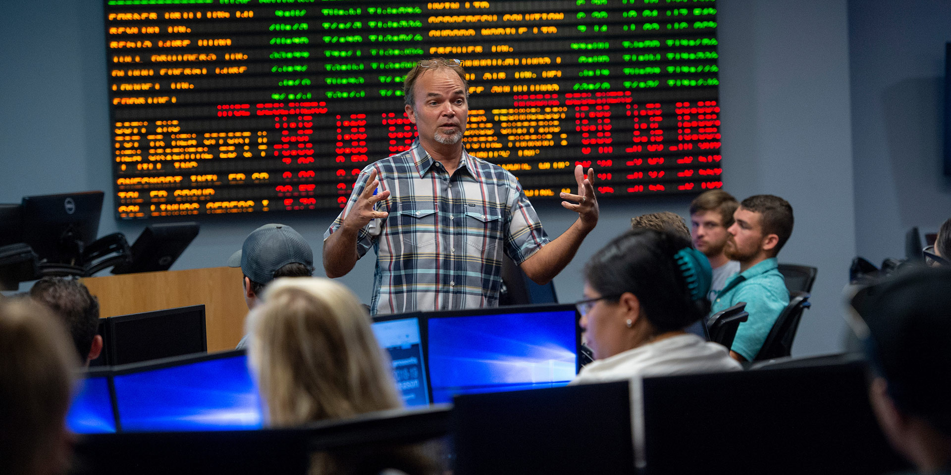 A professor gives a lecture in front of a large stock ticker board