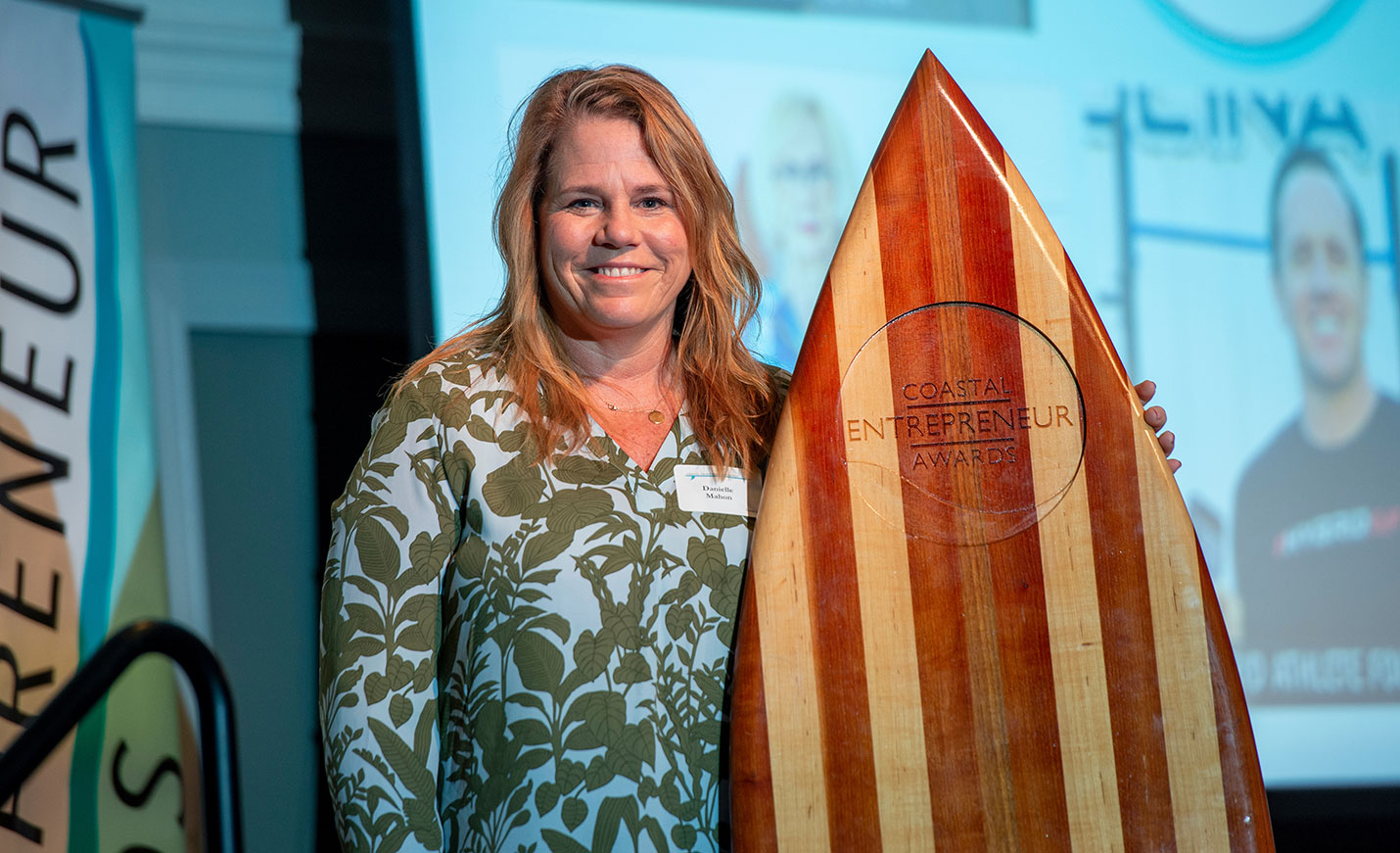 A woman stands next to a surfboard engraved with the Coastal Entrepreneurship Awards name and logo