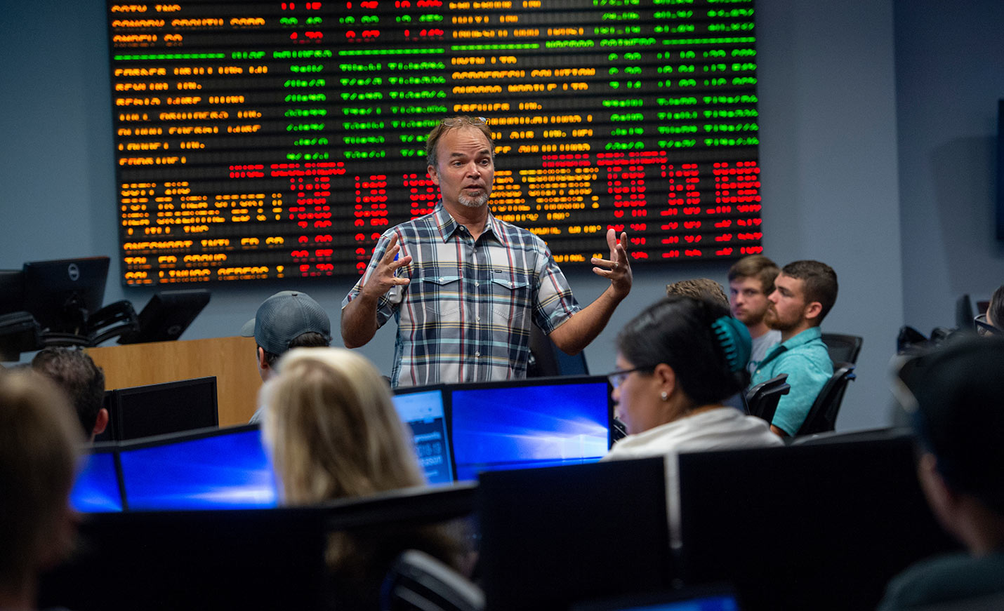A professor gives a lecture in front of a large group of people while standing in front of a stock ticker board