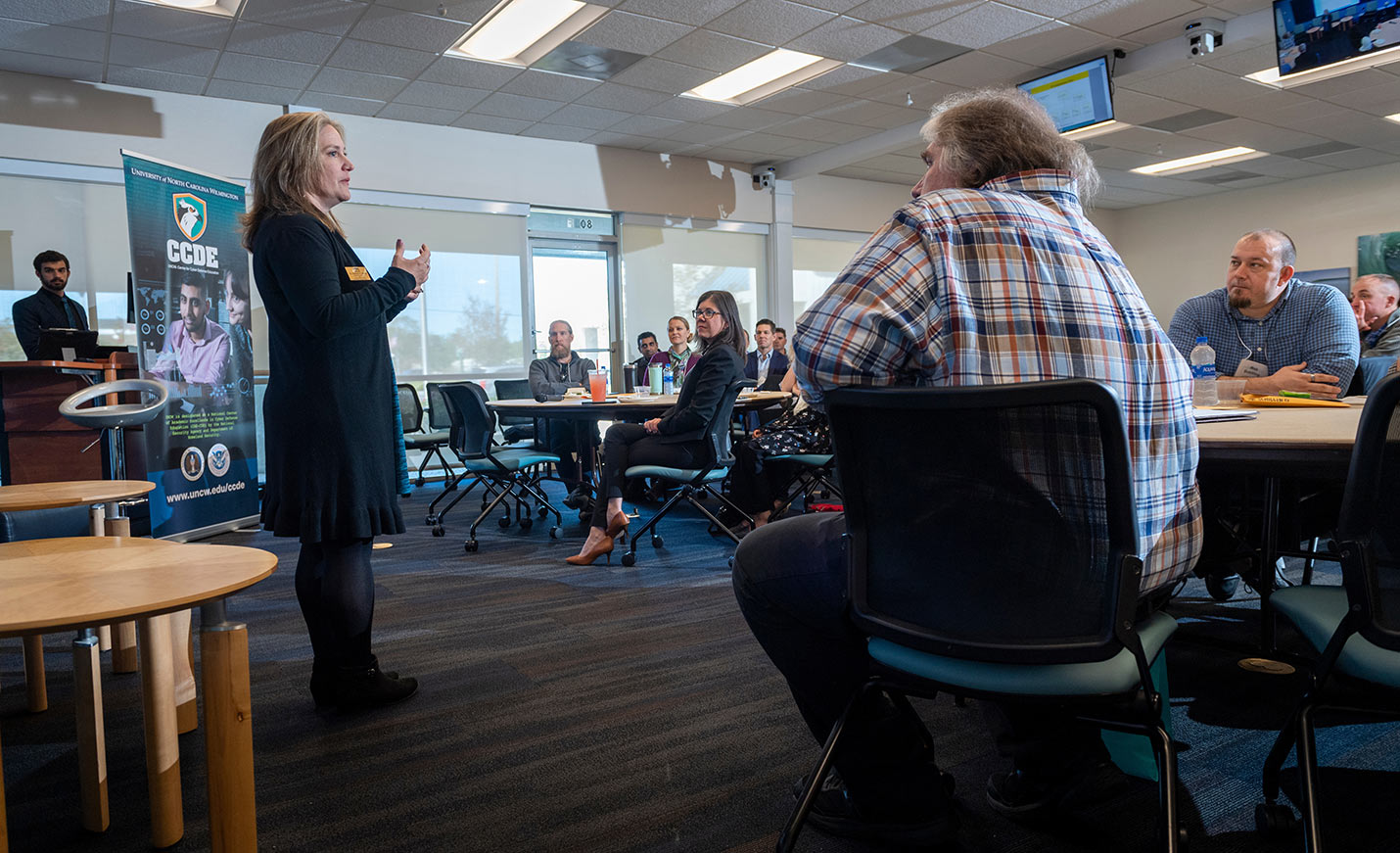 A speaker at a Center for Cyber Defense Education event talks to a group of people who are seated at tables