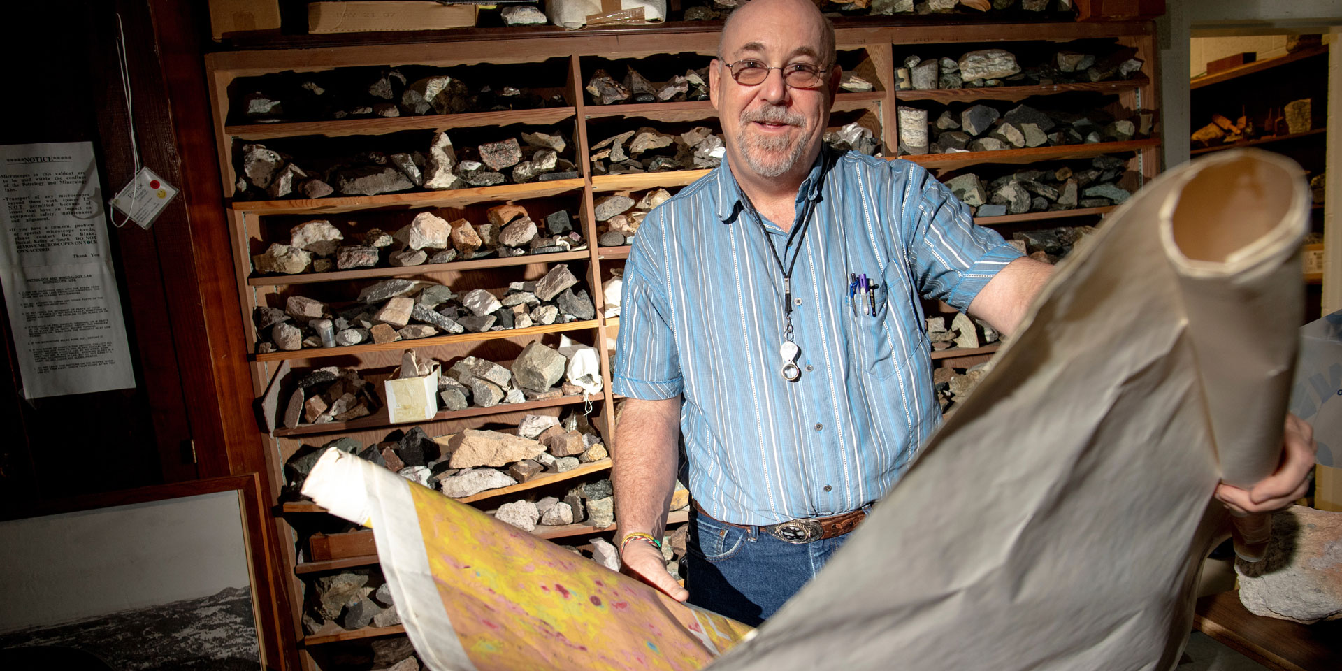 A professor stands holding a map in front of a shelf full of rocks 