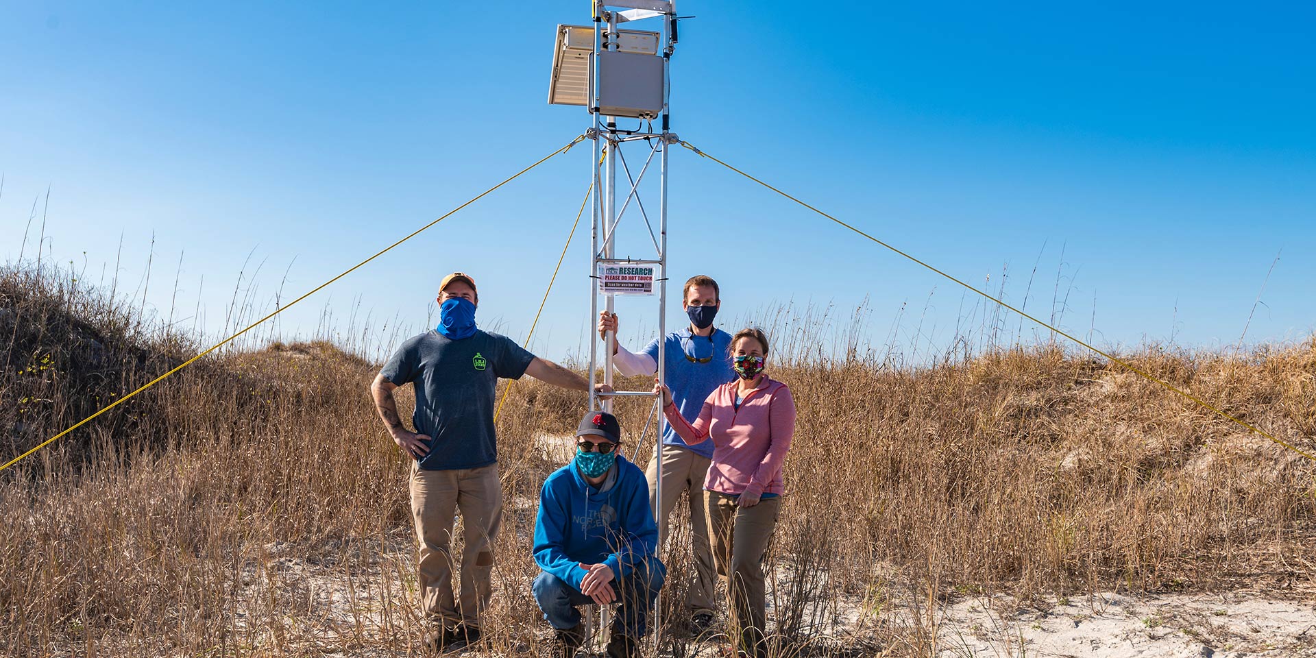 Four people stand next to a weather data tower on a dune